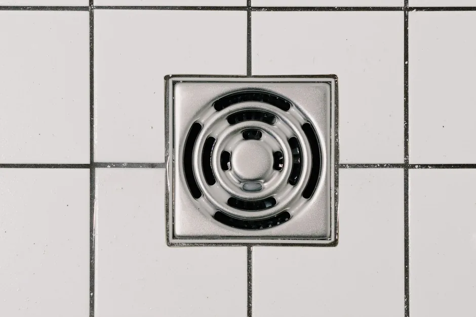 A square chrome floor waste grate set into white bathroom tiles