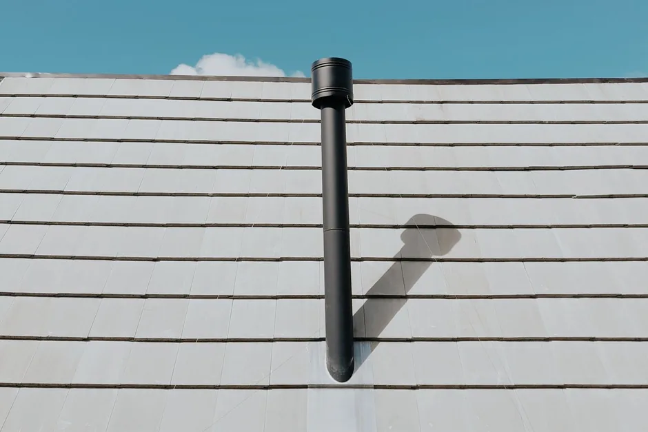 A black plumbing vent pipe rising through a slate roof against a blue sky