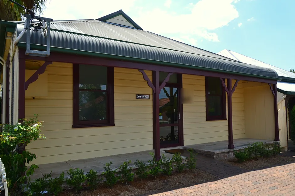 A single-storey yellow weatherboard cottage with a curved corrugated verandah roof, dark red posts and a downpipe at the front corner