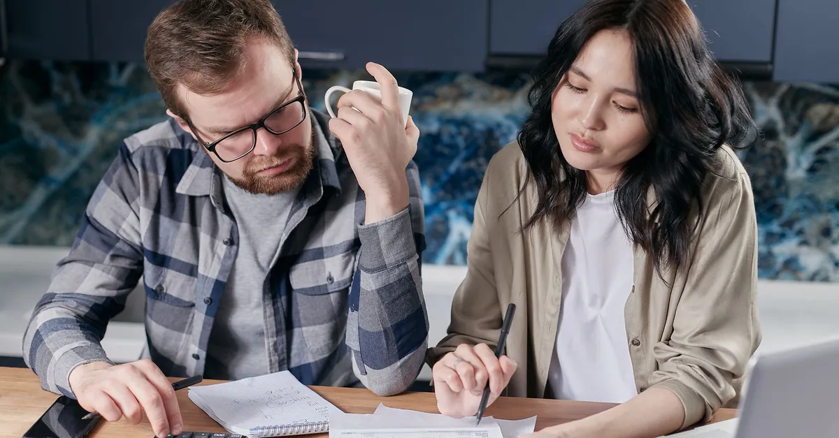 Two people comparing paperwork with a calculator at a kitchen table